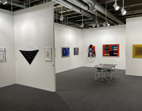 Inside small art exhibition booth with paintings on white walls, small table, and dark grey sustainable carpet floor